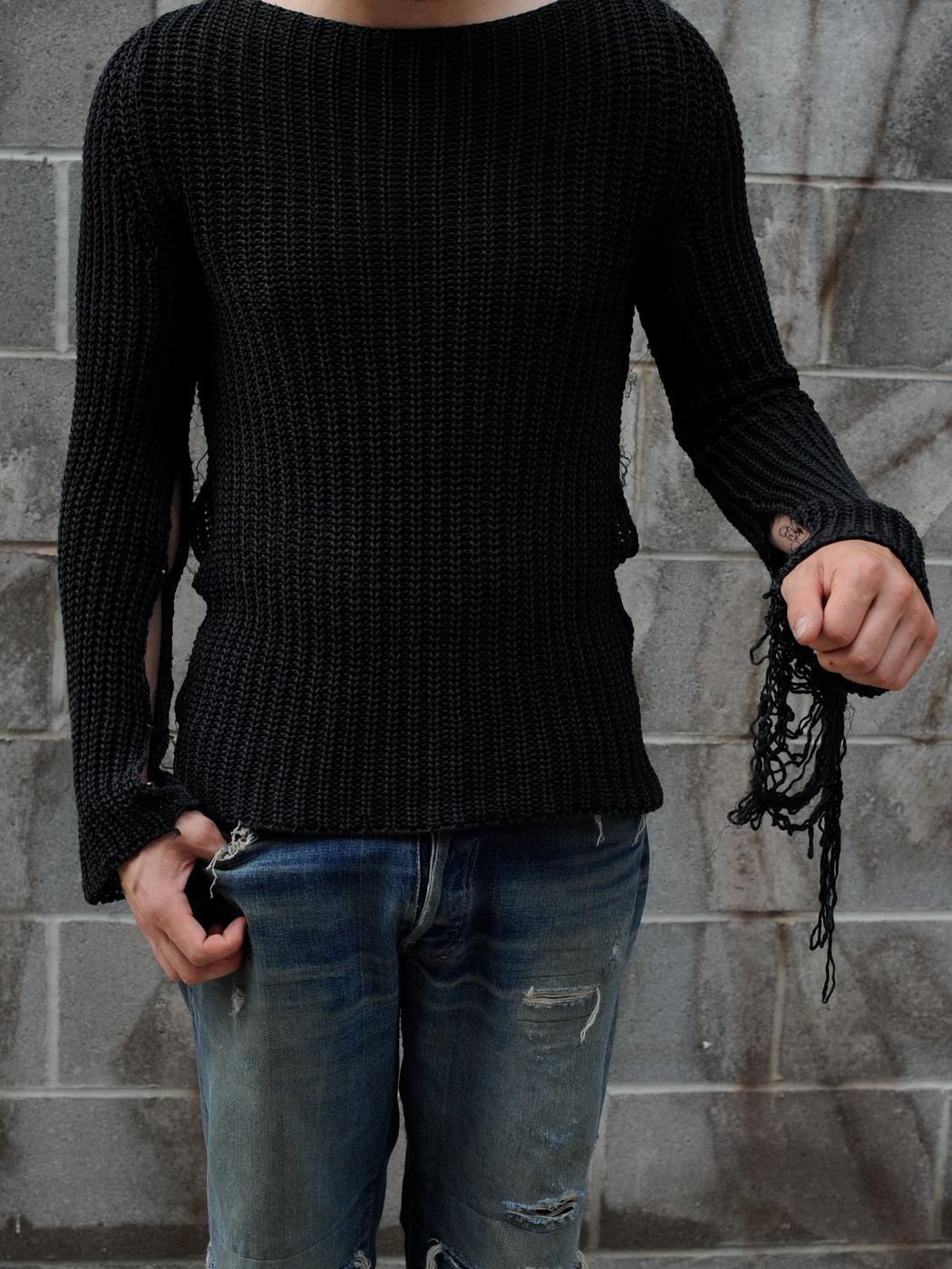 A black open knit sweater with a frayed cuff, worn against a grey block wall