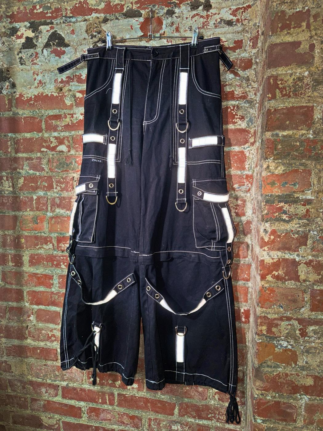 Black 3M Tripp NYC pants with reflective straps and D rings, hung against a painted brick wall